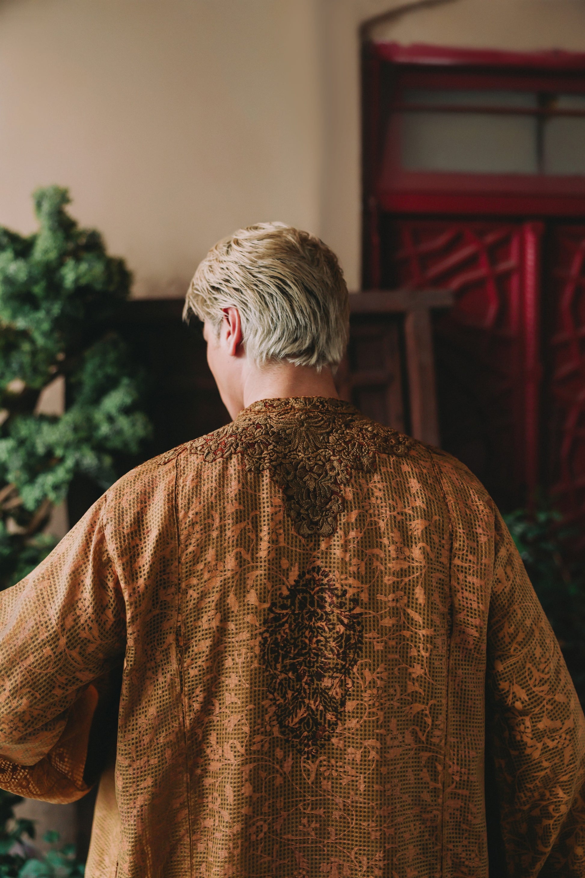 Person wearing a textured brown garment standing in a room with a red door and greenery.