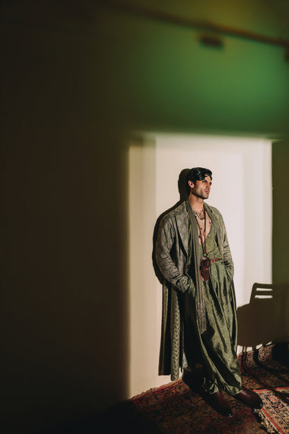 Man in traditional green attire standing in a room with a white wall and a chair.