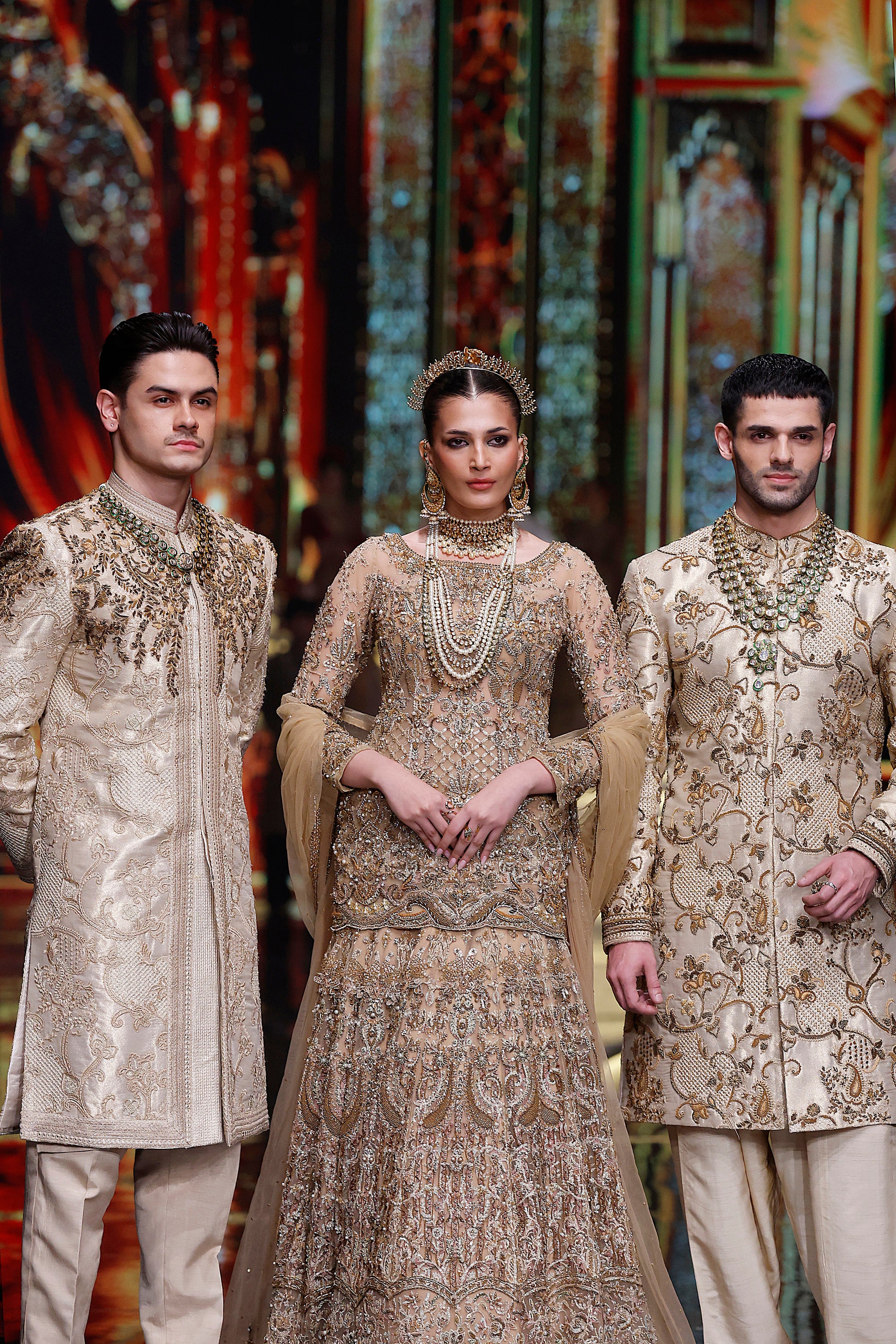 Three individuals in traditional embroidered outfits standing against a colorful, patterned background.