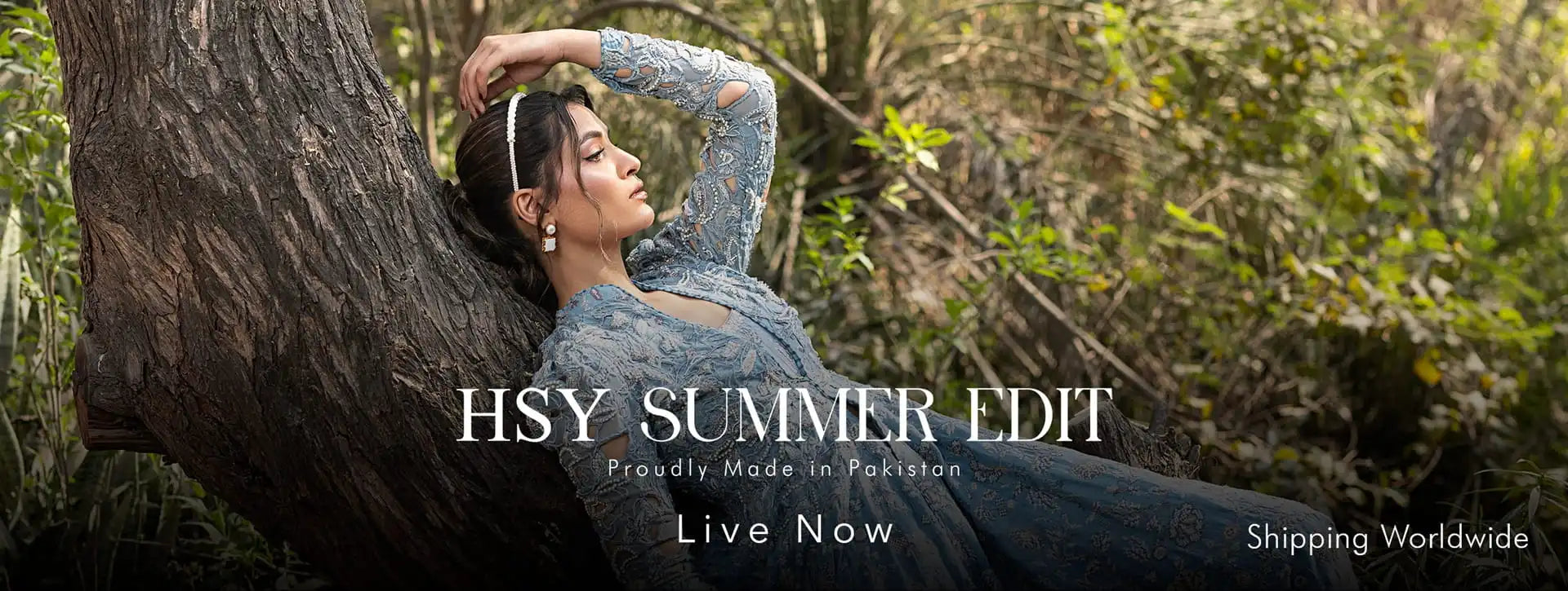 A woman reclines against a large tree trunk in a wooded outdoor setting, wearing a sheer long-sleeve blue dress. White text overlays read 'HSY SUMMER EDIT', 'Proudly Made in Pakistan', 'Live Now', and 'Shipping Worldwide'.
