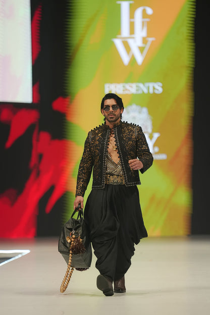 Model walking on a runway wearing a black outfit with intricate designs, holding a bag, with a colorful 'IFW' branded backdrop.