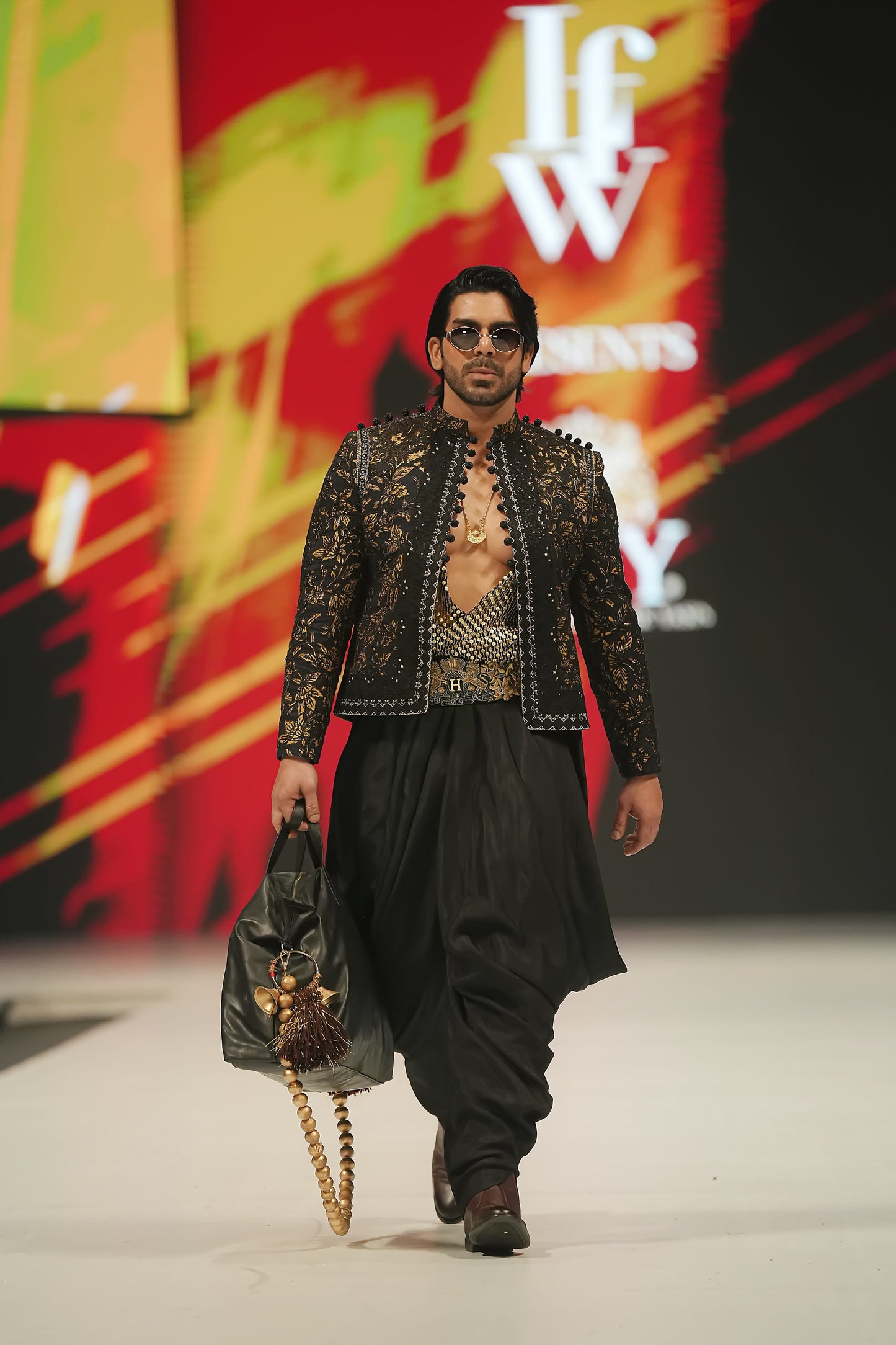 Model walking on a runway wearing a patterned jacket and black pants, with a colorful 'Ifw' backdrop.