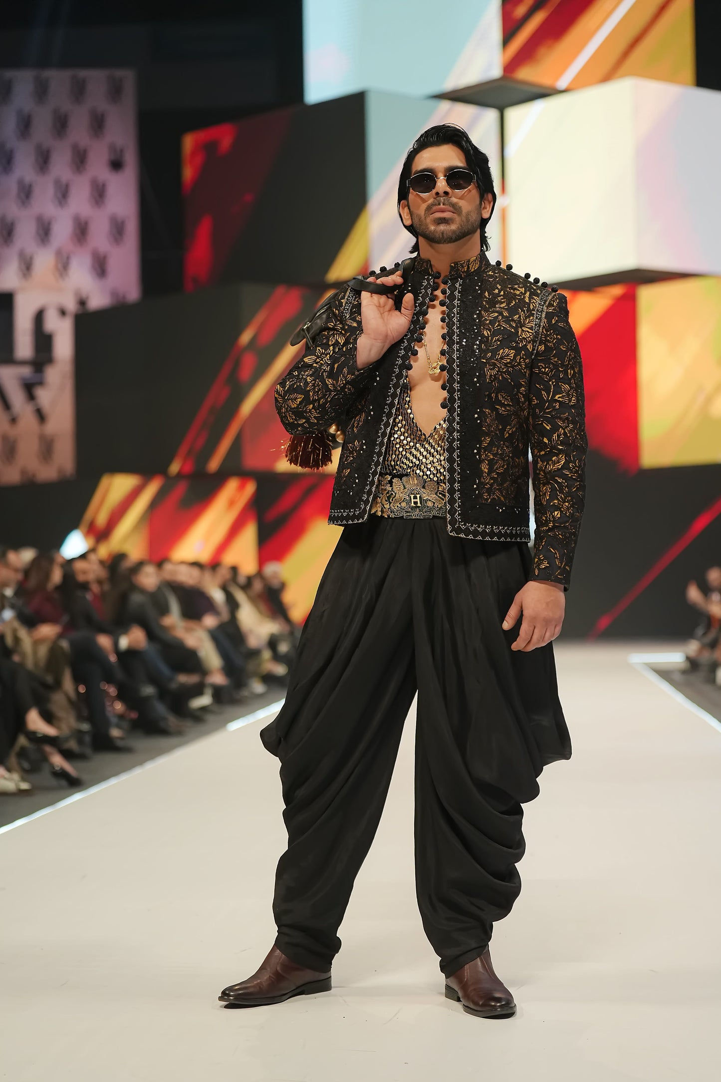 Man modeling a traditional outfit on a fashion show runway with a colorful background.