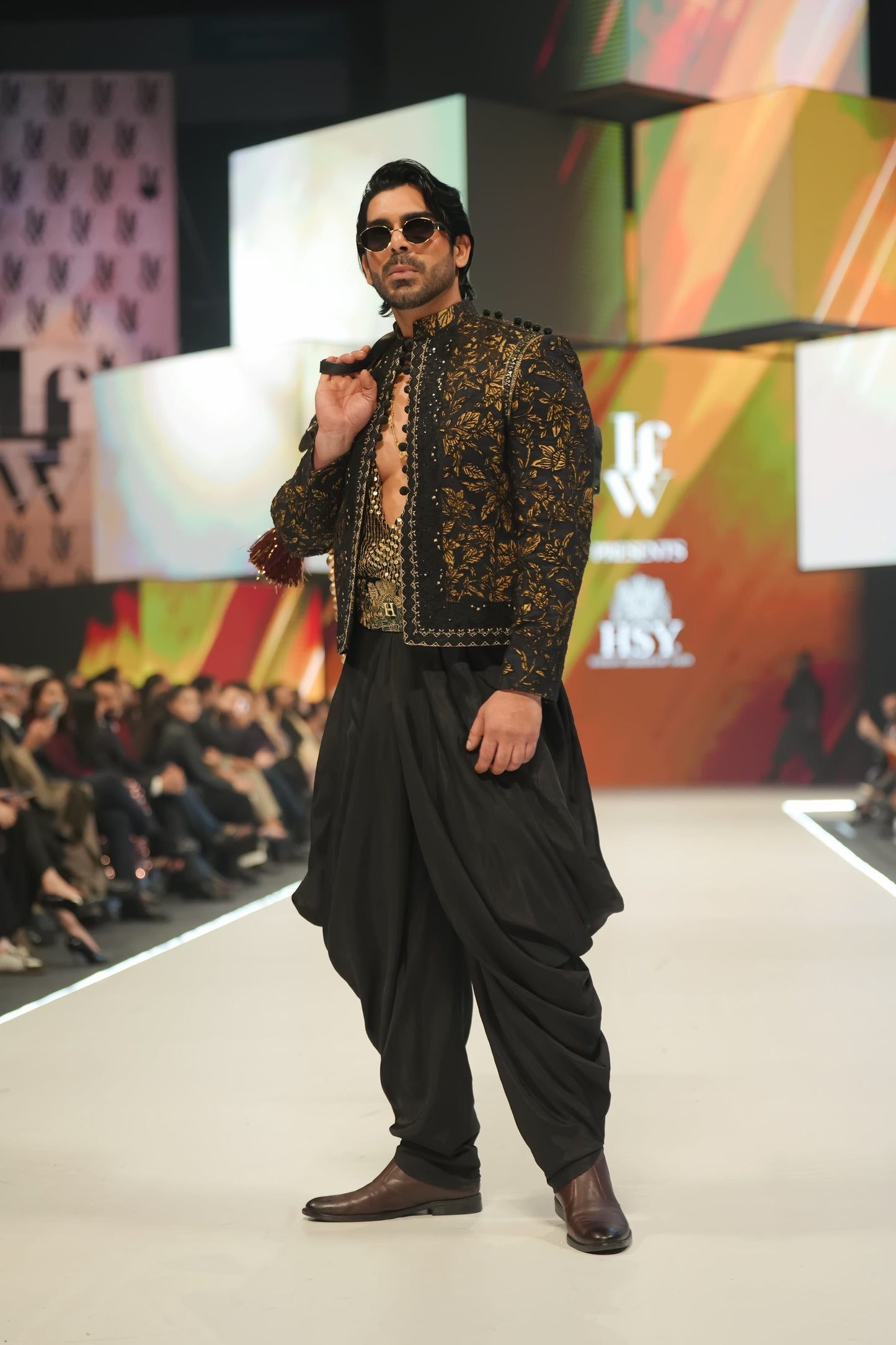 Model walking on a fashion show runway wearing a black and gold outfit.