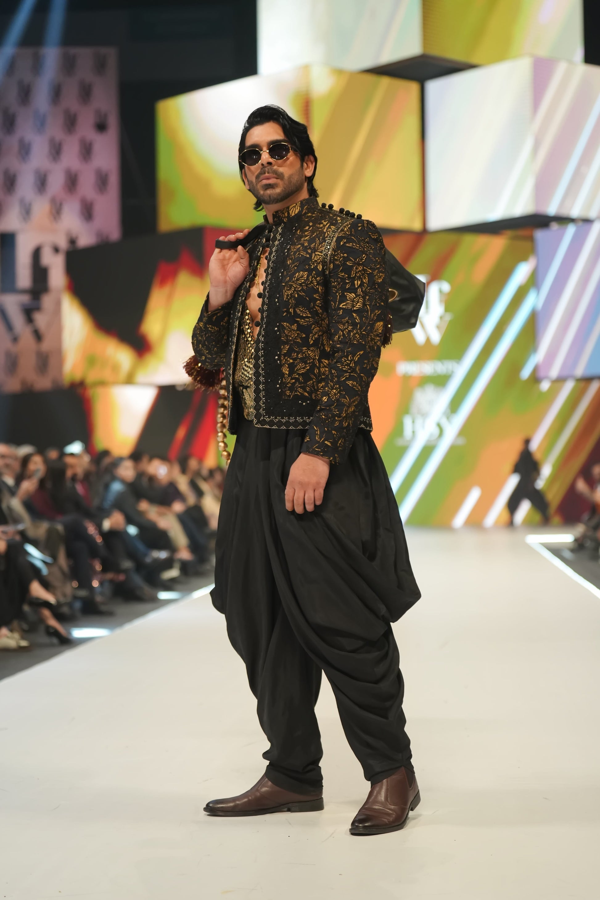 Man walking on a fashion show runway wearing a patterned jacket and black pants.