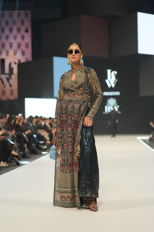 Model walking on a fashion show runway wearing a detailed outfit with sunglasses and earrings.