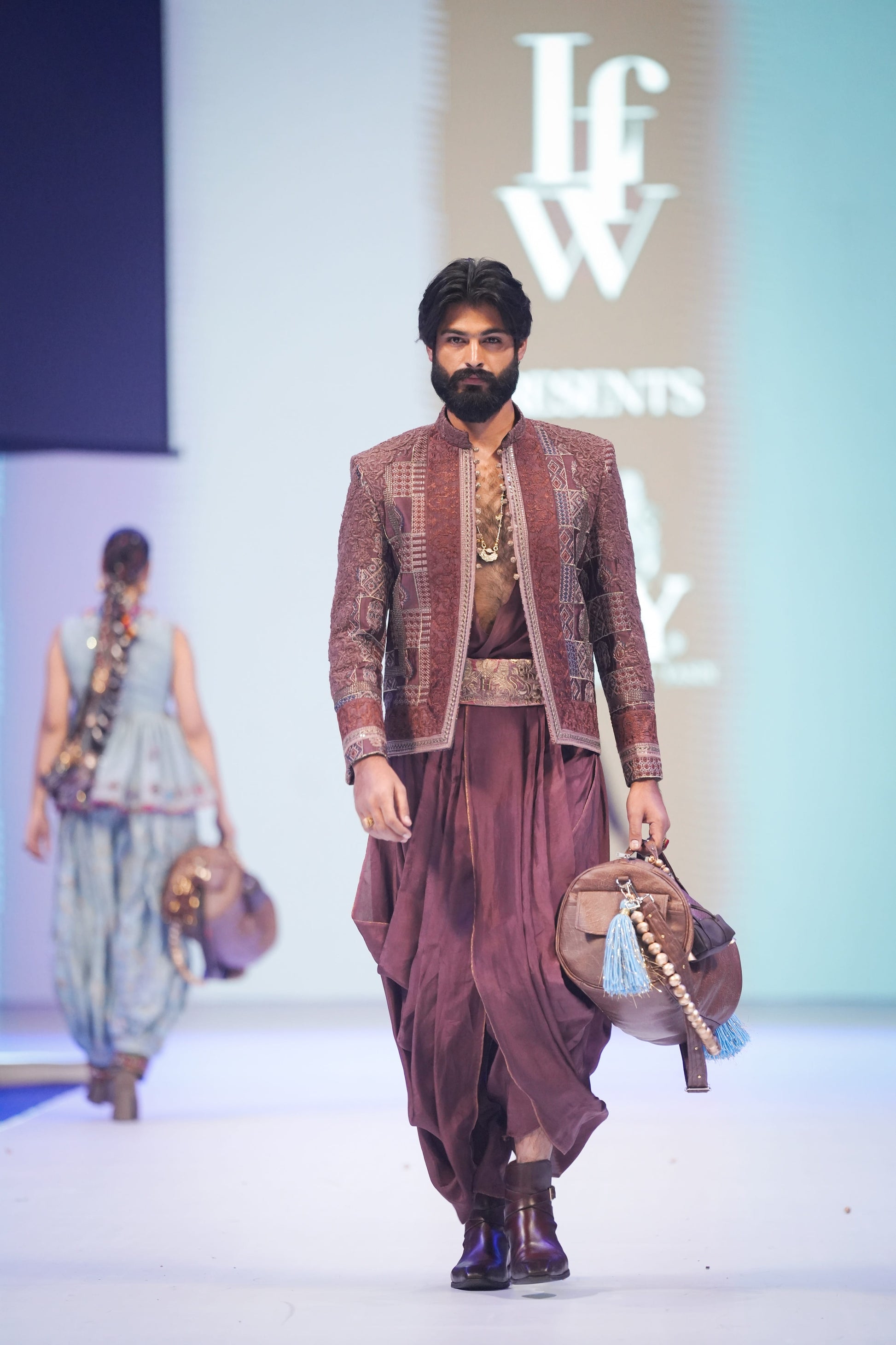 Man in a maroon traditional outfit walking on a fashion show runway with 'IFW' branding in the background.