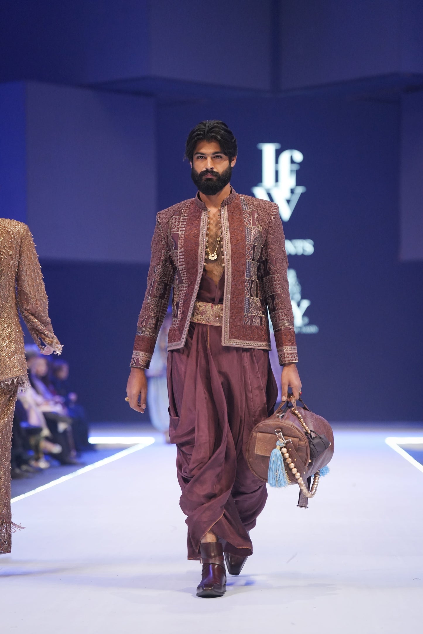 Man in traditional maroon outfit walking on a runway with 'lfw' branding in the background