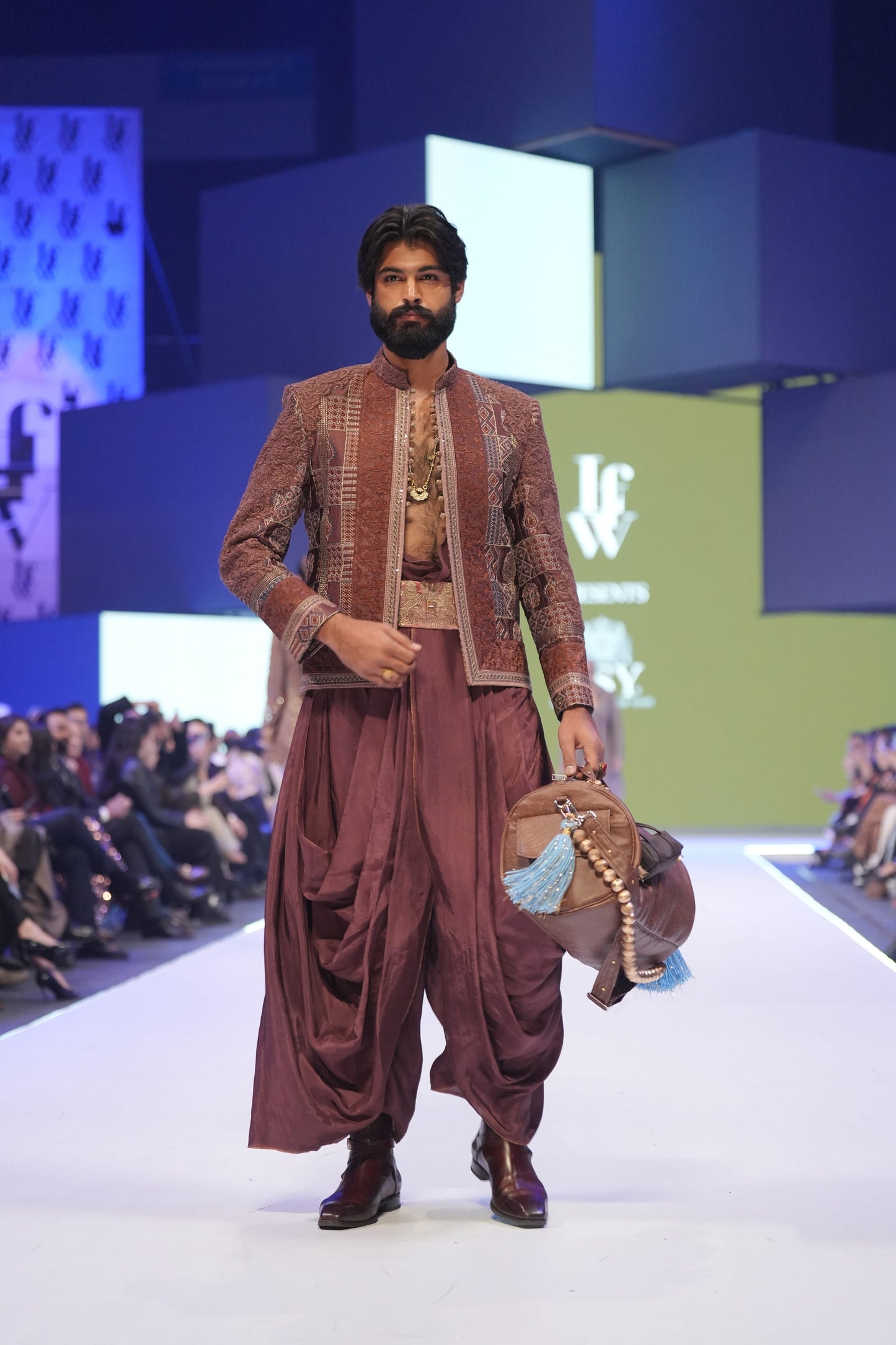 Man in traditional maroon outfit with gold details on a fashion show runway.