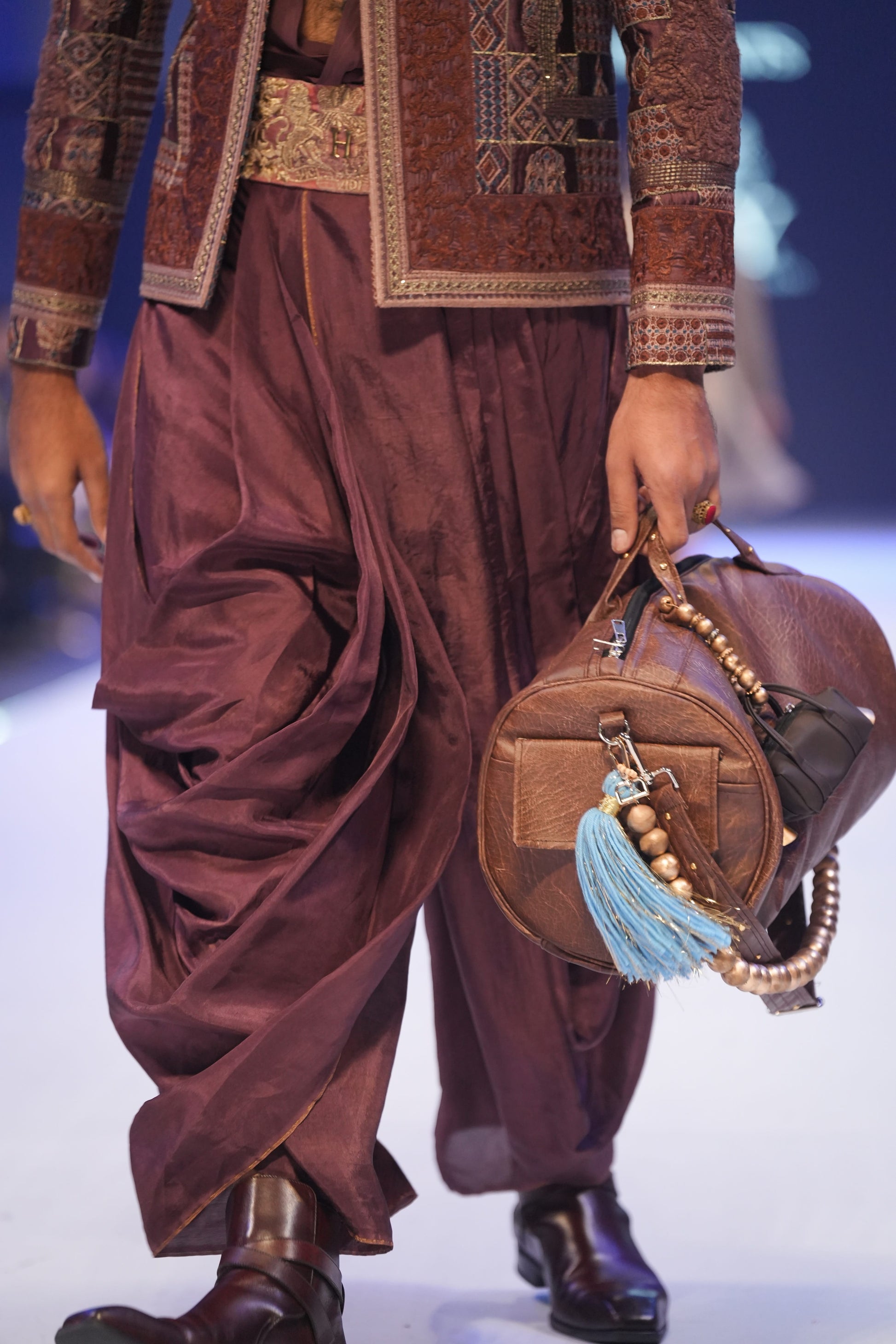 Person wearing traditional maroon attire with a matching handbag on a runway.