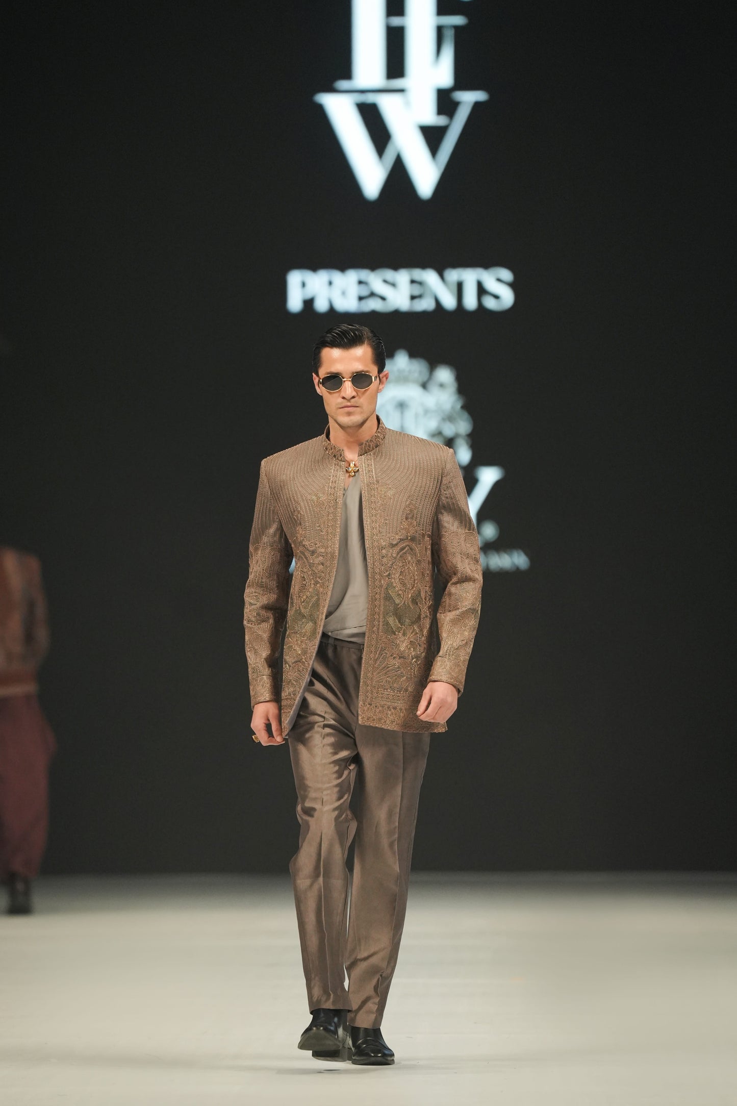 Man walking on a fashion show runway wearing a brown outfit with sunglasses, against a backdrop featuring a logo and text.