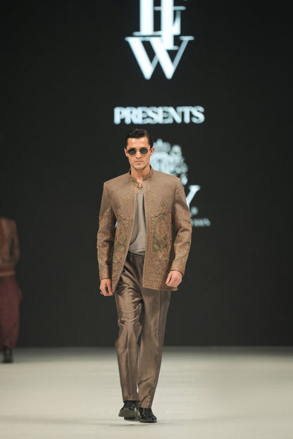 Man walking on a fashion show runway wearing a brown outfit with sunglasses, against a backdrop featuring a logo and text.