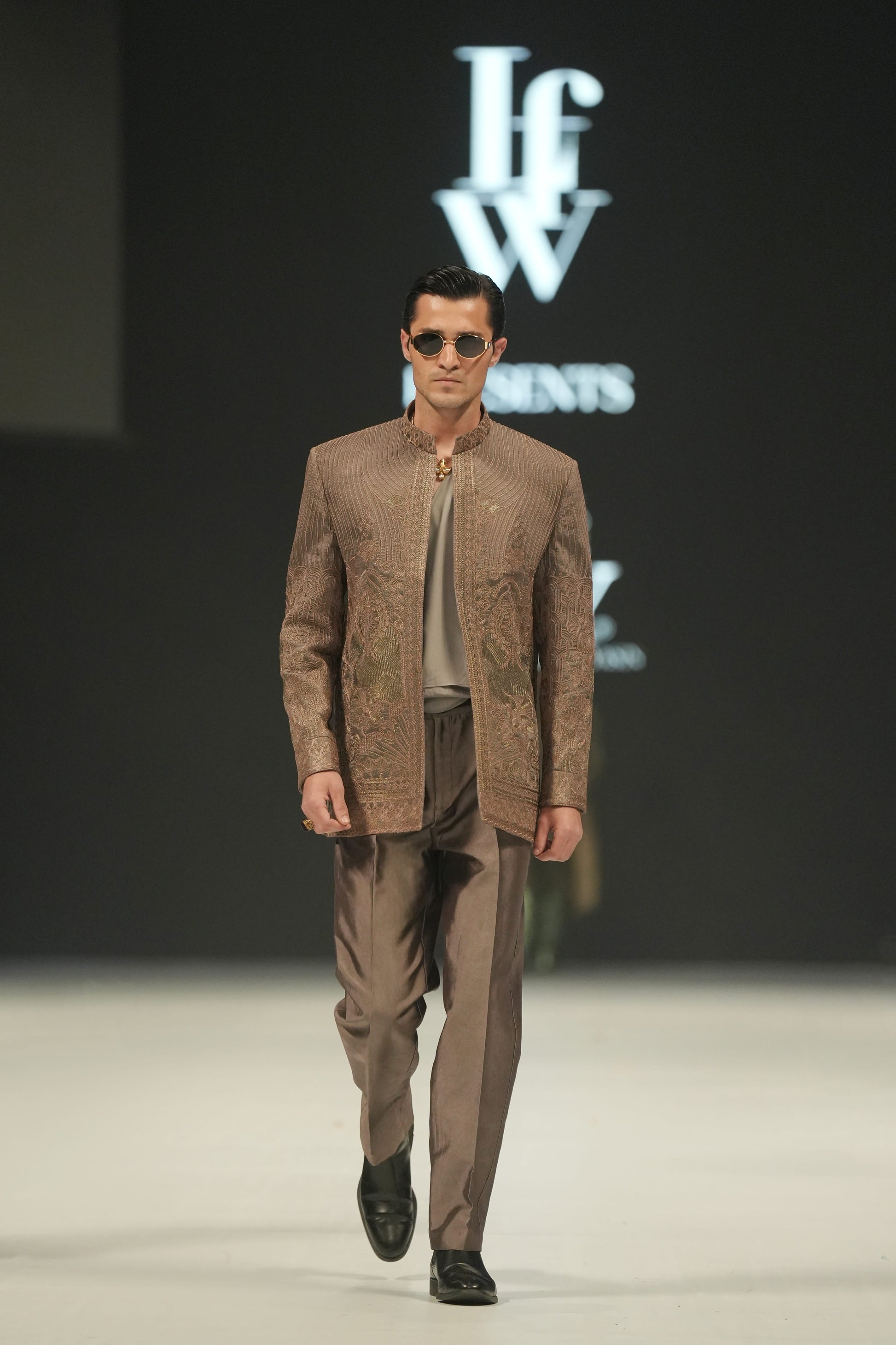 Man walking on a runway wearing a brown suit with sunglasses, against a dark background with 'lFW' branding.