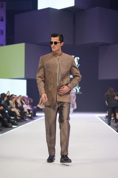 Man in a brown suit walking on a fashion show runway with a blurred audience in the background.