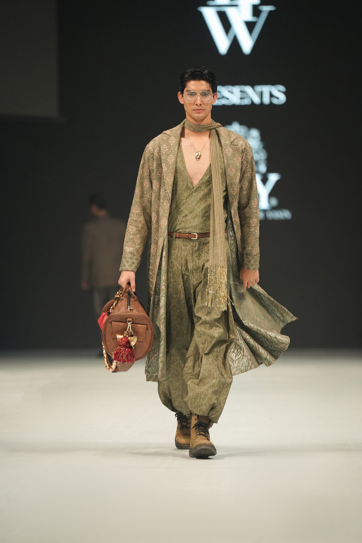 Model walking on a runway wearing a green outfit with a bag, with 'LFW' and 'HSY' logos in the background.