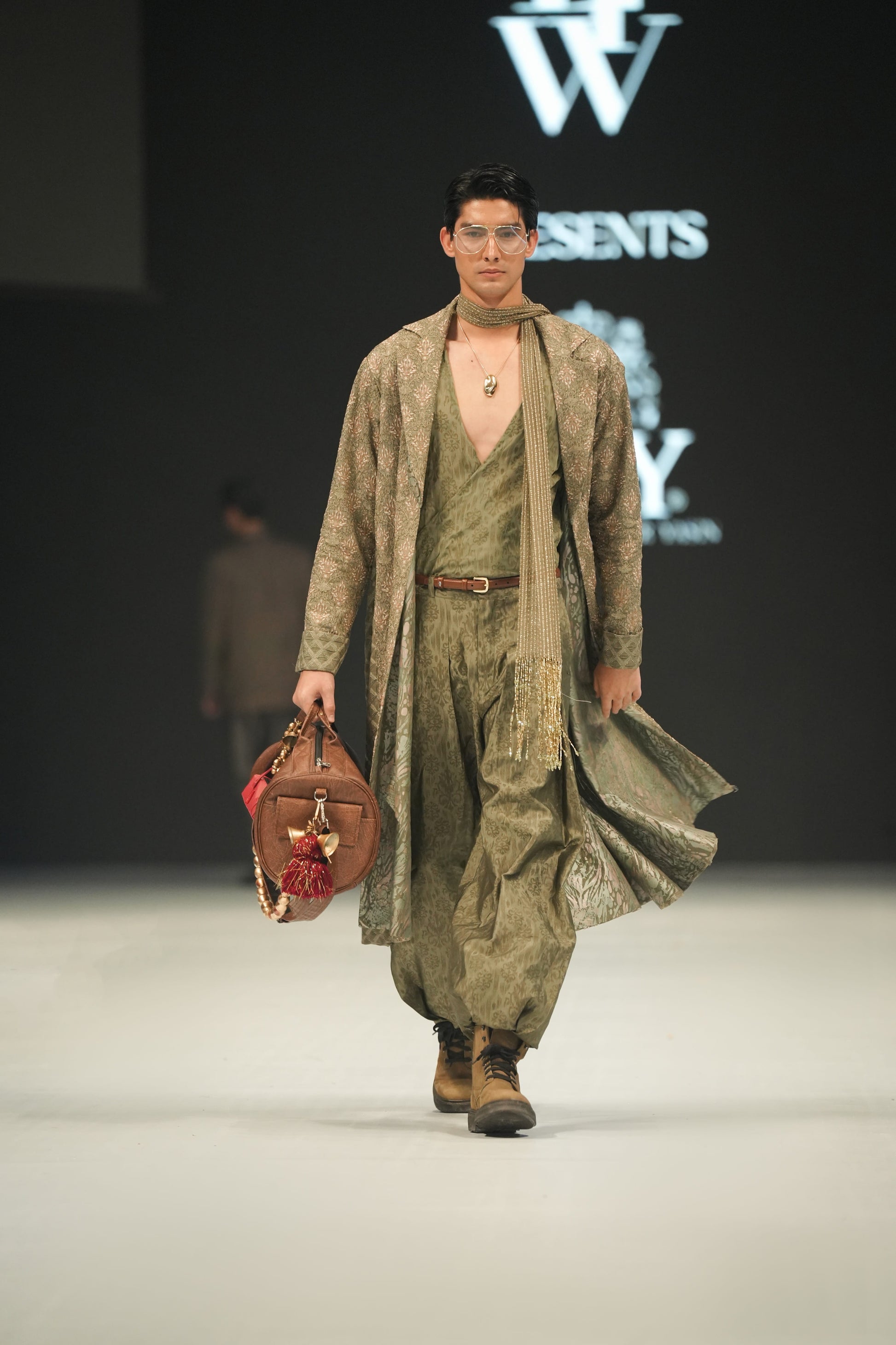 Model walking on a runway wearing a green outfit with a bag, with 'LFW' and 'HSY' logos in the background.
