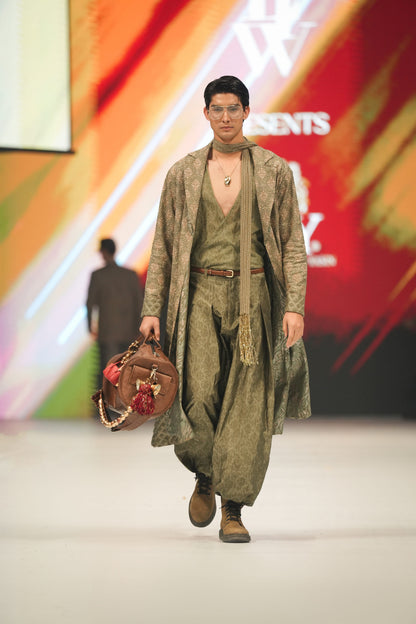 Model walking on a fashion show runway wearing a green patterned outfit with a brown bag.