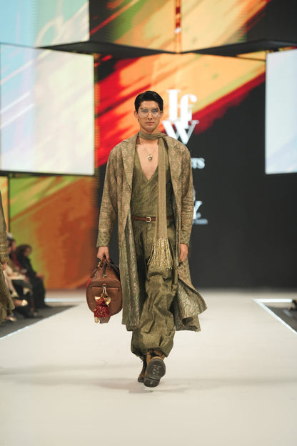 Model walking on a fashion show runway wearing a long, patterned coat and holding a bag.