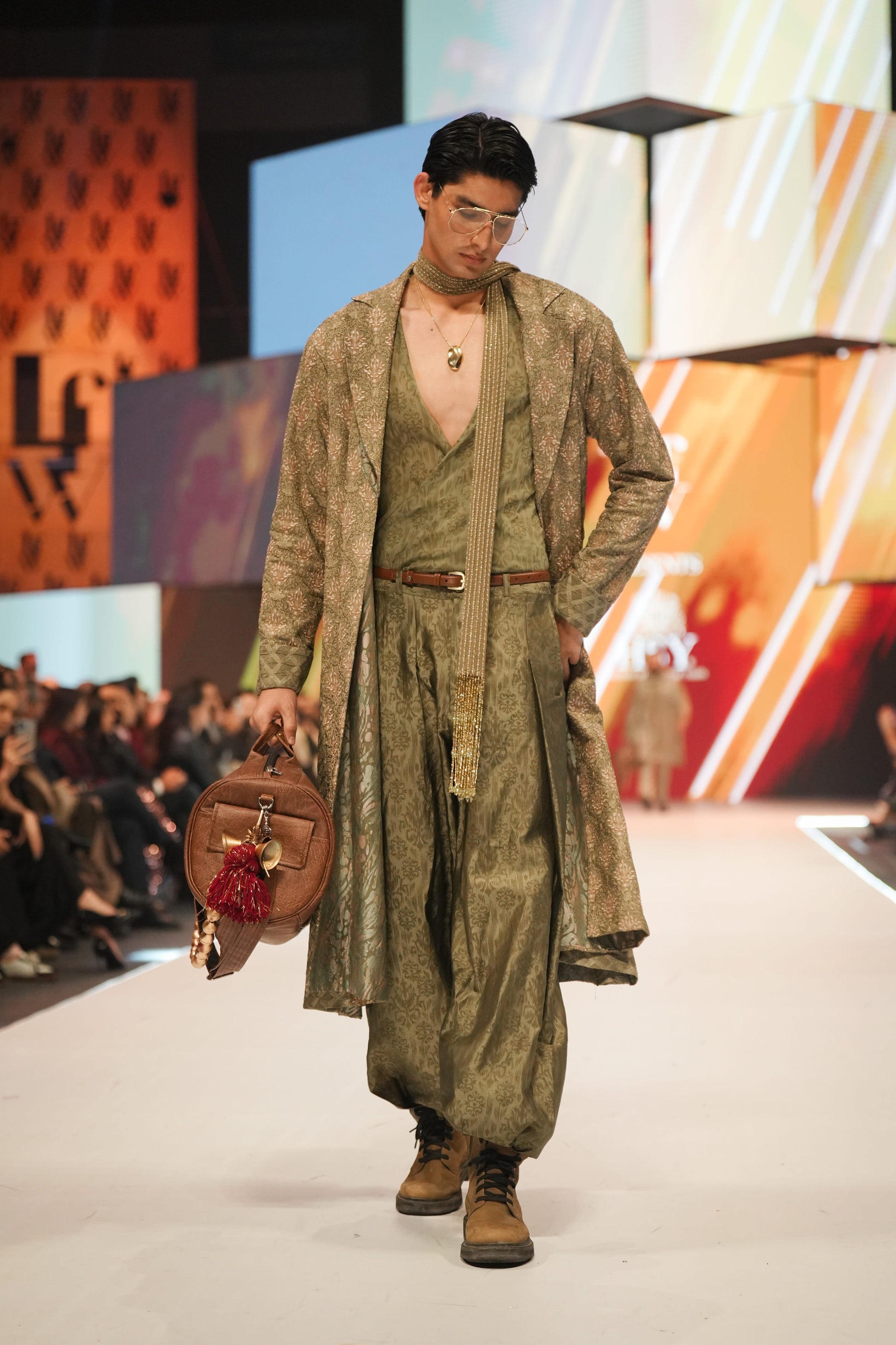 Model walking on a runway wearing a long green coat and holding a bag, with a colorful backdrop.