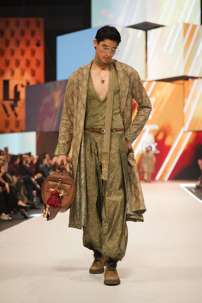 Model walking on a runway wearing a long green coat and holding a bag, with a colorful backdrop.