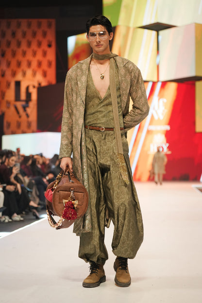 Model walking on a fashion show runway wearing a green outfit with a brown bag.