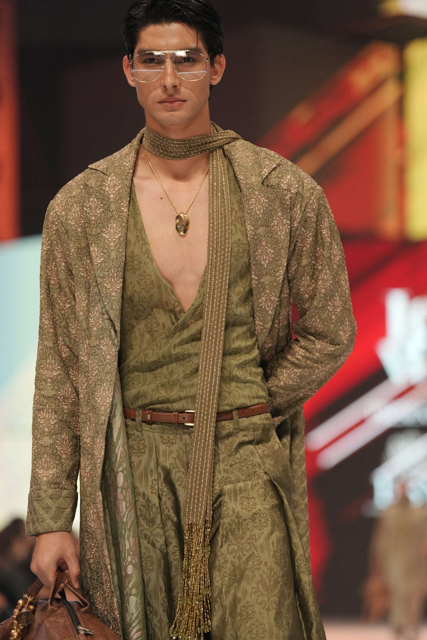 Man modeling a green outfit with a patterned shawl on a runway.