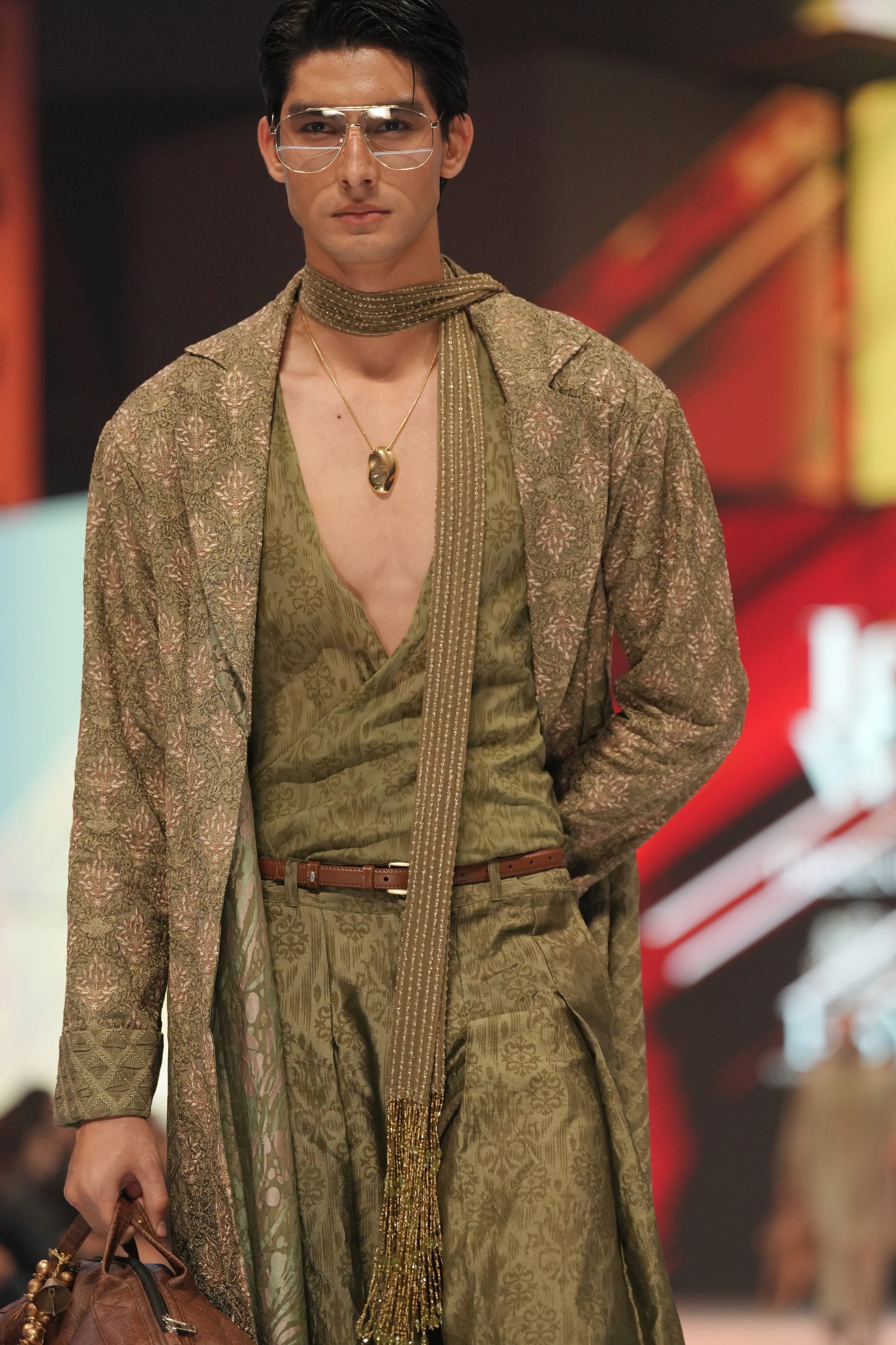 Man modeling a green outfit with a patterned shawl on a runway.