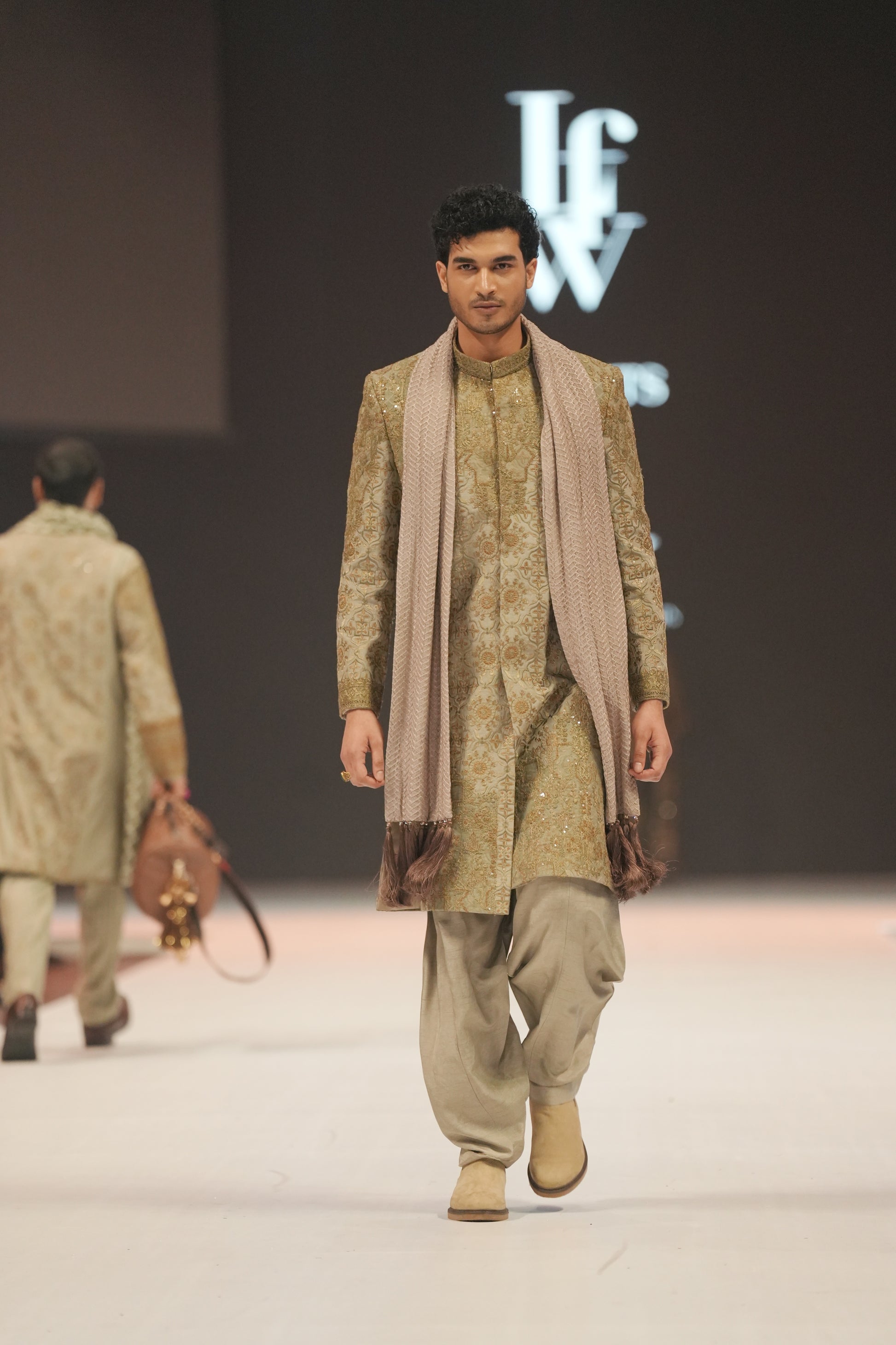 Man walking on a fashion runway wearing a traditional outfit with a brand logo in the background