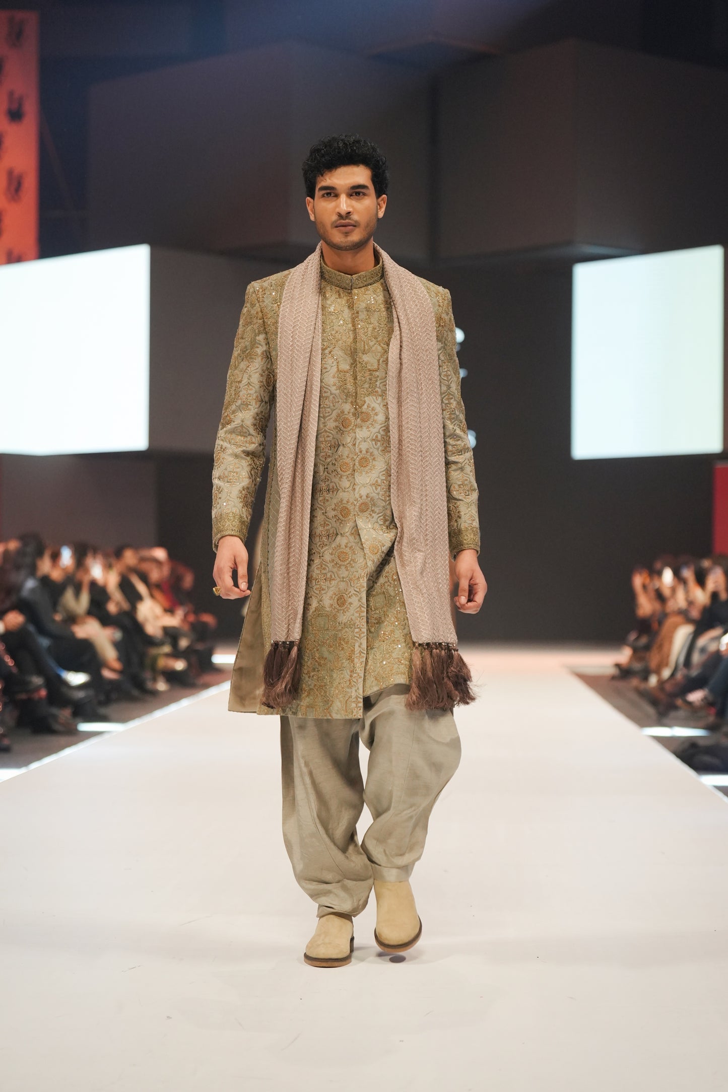 Man walking on a fashion show runway wearing a traditional outfit with a scarf.