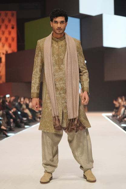 Man walking on a fashion show runway wearing a traditional outfit with a long scarf.