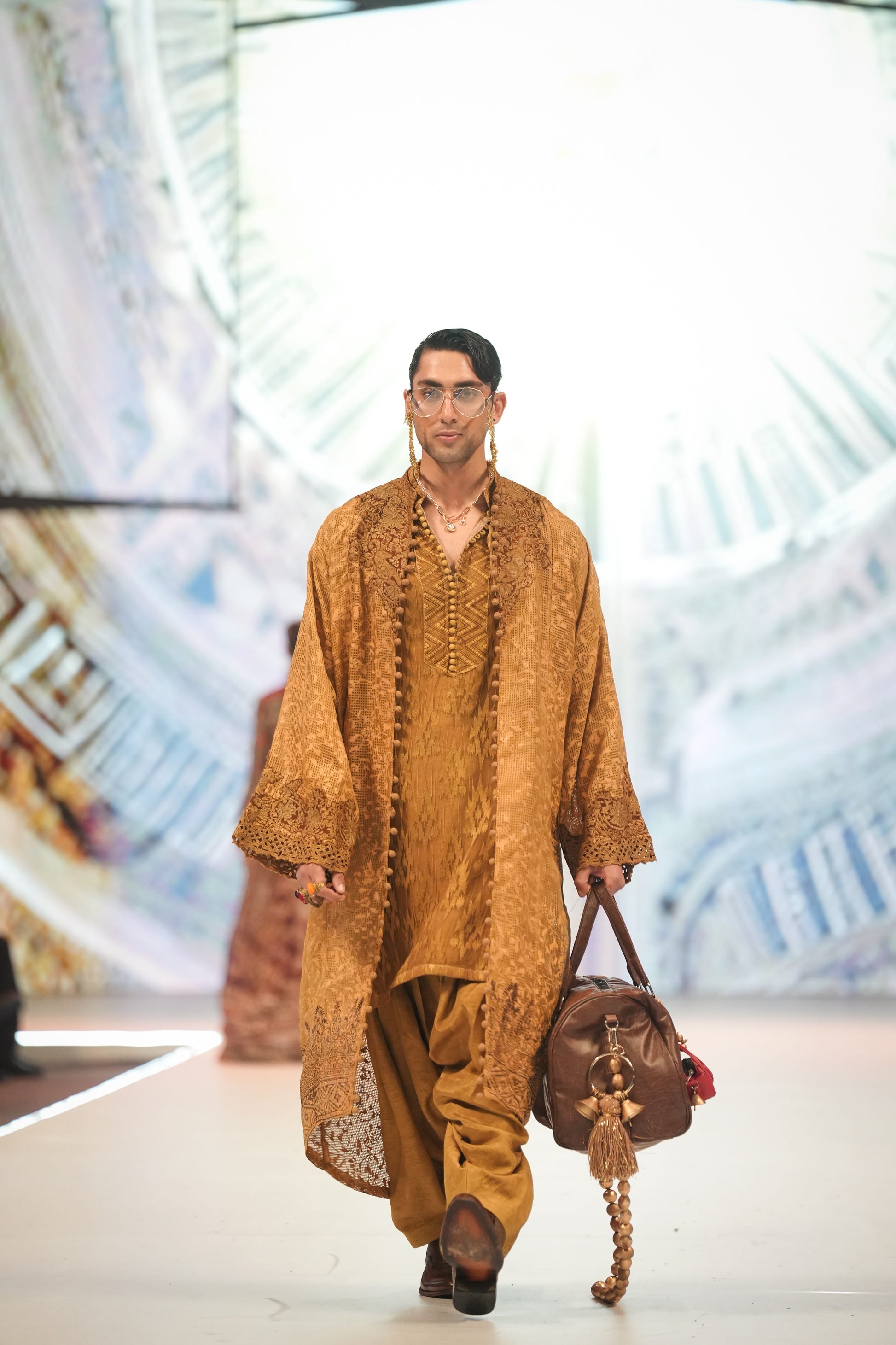 Man walking on a runway wearing a traditional outfit with a brown bag.