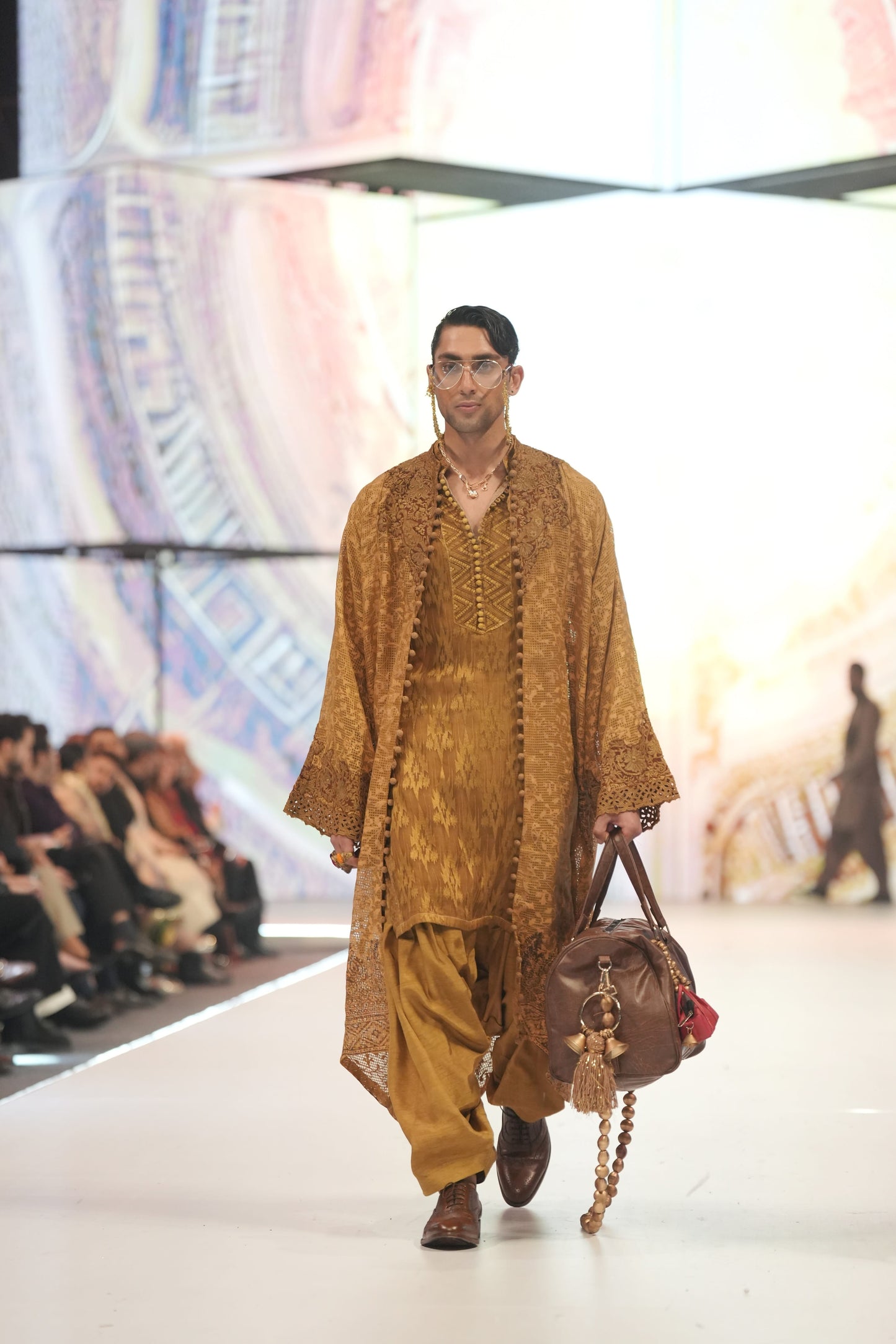 Man walking on a fashion show runway wearing a brown outfit with a large bag.