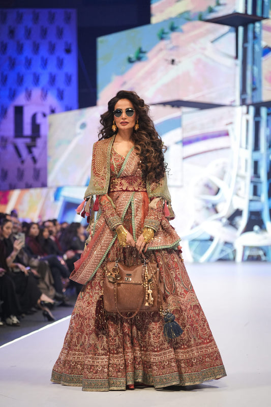 Model walking on a fashion show runway wearing a traditional outfit with a handbag.